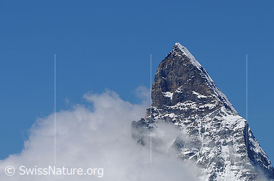 Foto: Gipfel des Matterhorns. Über den von rechts aufsteigenden Nordostgrat (NE-Grat, Hörnligrat) führt die Normalroute von der Schweizer Seite. Gut zu sehen sind die Schulter und das Dach. In der Fallinie des Gipfels ist der oberste Teil der Ostwand (E-Wand) zu sehen..